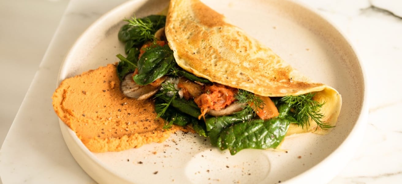 Golden omelette filled with fresh spinach, herbs, and vegetables, served with a spread of orange hummus on a beige plate.